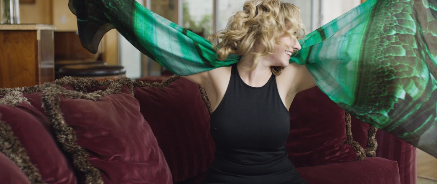 A woman with curly blonde hair sitting on a red velvet couch, smiling and holding a green and black patterned fabric above her head.