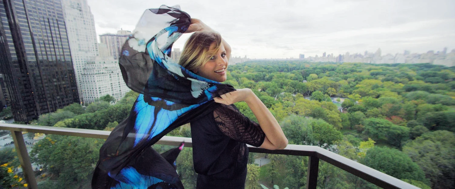 Woman standing on a balcony overlooking a lush green park with skyscrapers in the background, holding a colorful sheer scarf around her head and shoulders.