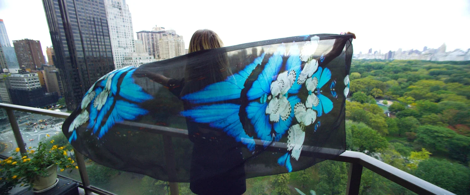 Person holding a large sheer scarf with a blue butterfly design on a balcony overlooking a cityscape and green park.