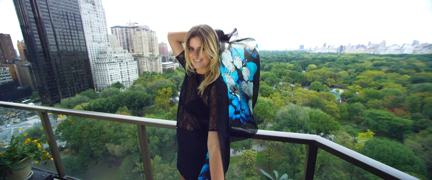 Woman with blond hair in a black lace top and black skirt standing on balcony overlooking city park with tall buildings and trees.