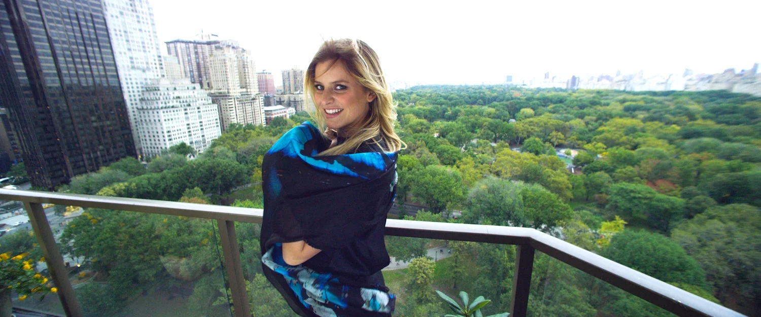 A woman with blonde hair smiling on a balcony overlooking a city and a large green park