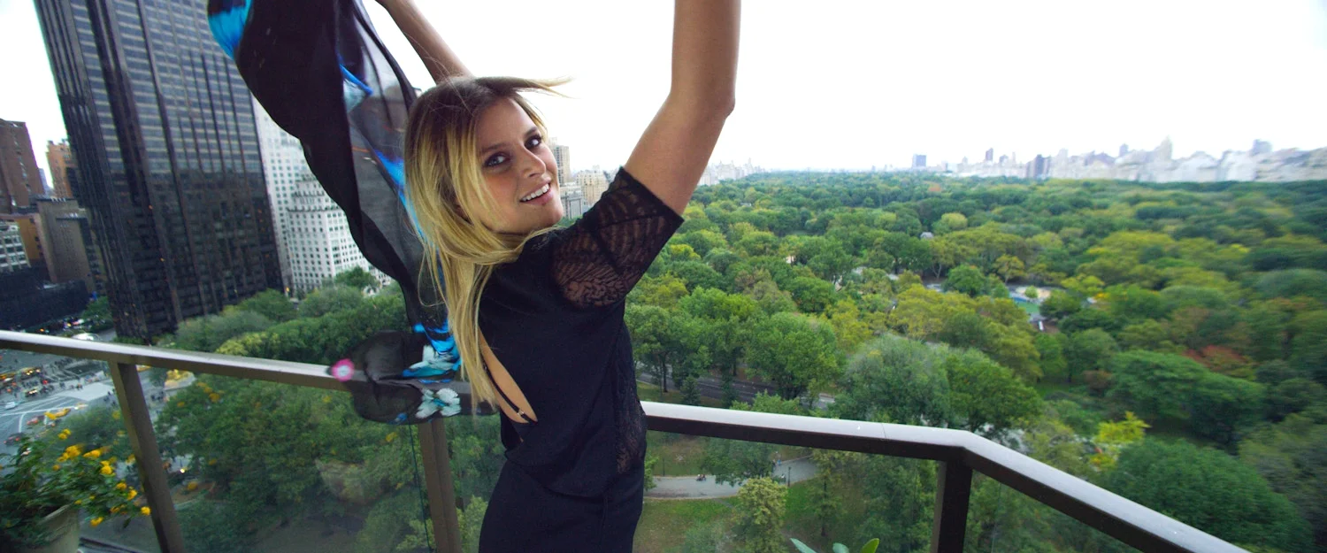 A woman with long blond hair, wearing a black lace top, holding a colorful jacket, standing on a balcony overlooking a city park with tall buildings and green trees.