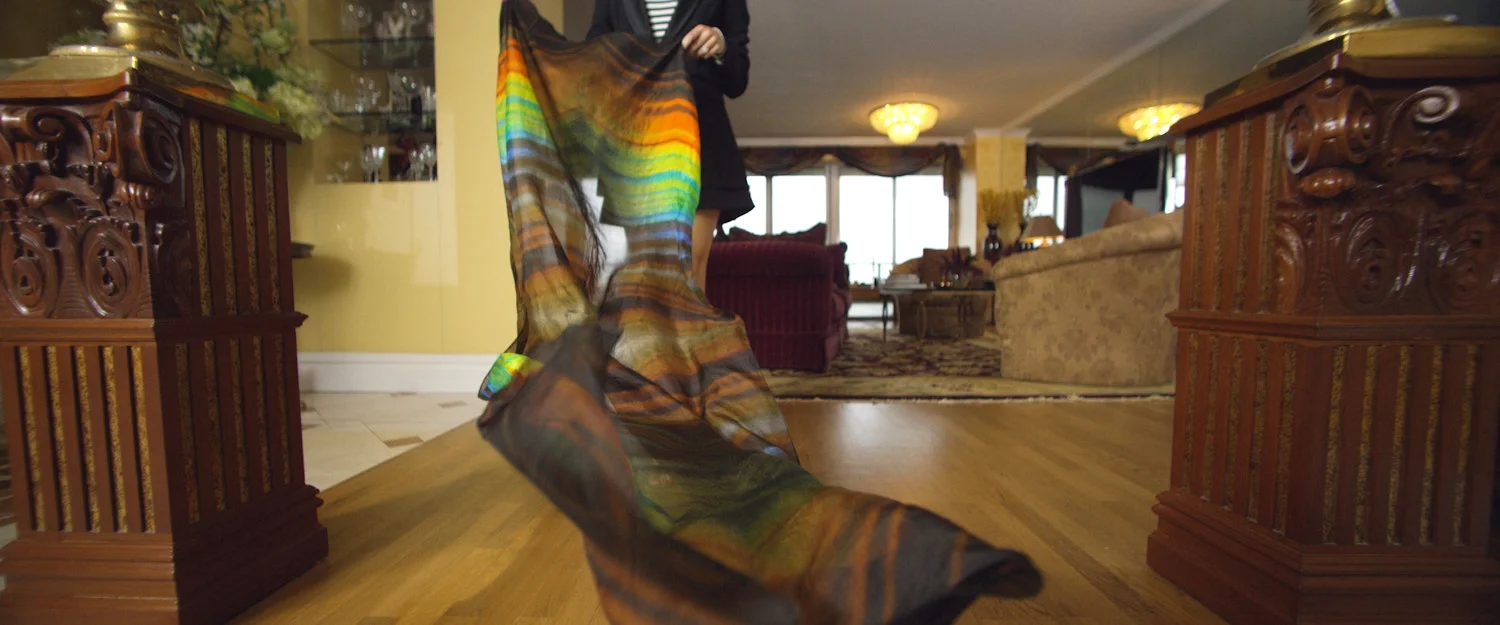 Person holding a colorful, flowing silk scarf indoors with wooden furniture and a living room in the background.
