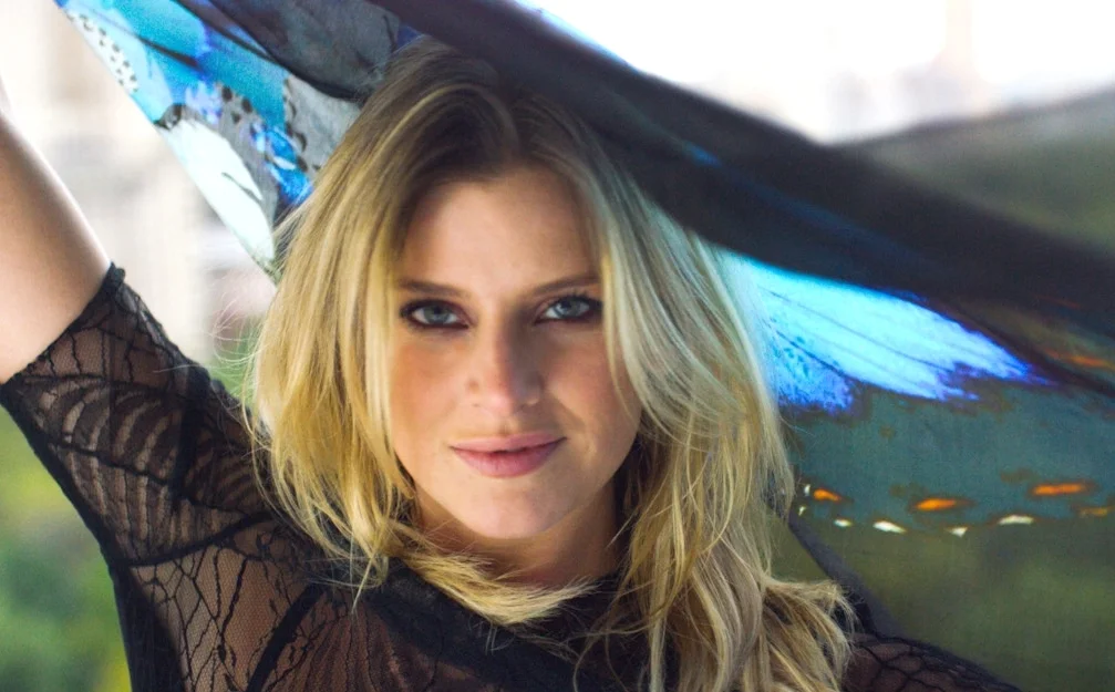A woman with blonde hair and fair skin smiling, wearing a black lace top, holding a colorful fabric above her head outdoors.