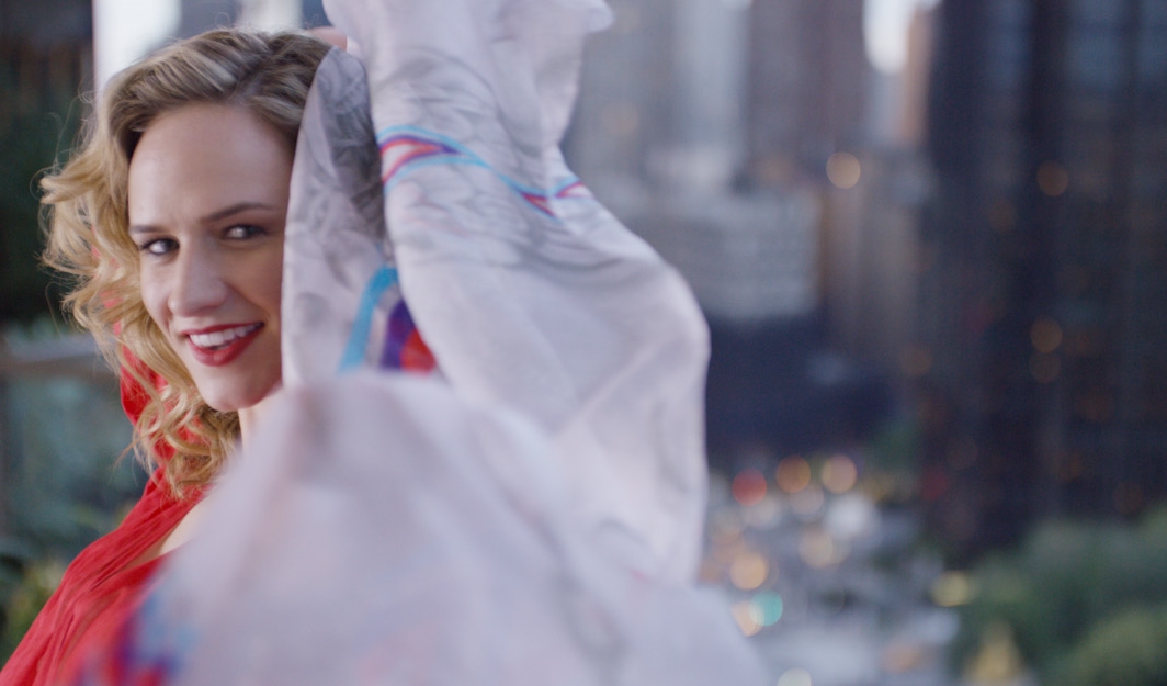 Woman with curly blonde hair wearing a red top, smiling and holding a plastic bag outdoors in an urban setting.