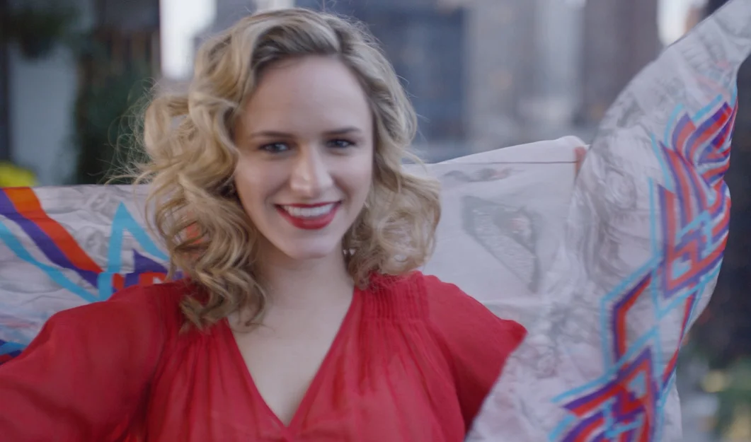A smiling woman with curly blonde hair wearing a red top, holding a colorful, patterned shawl or fabric behind her shoulders.