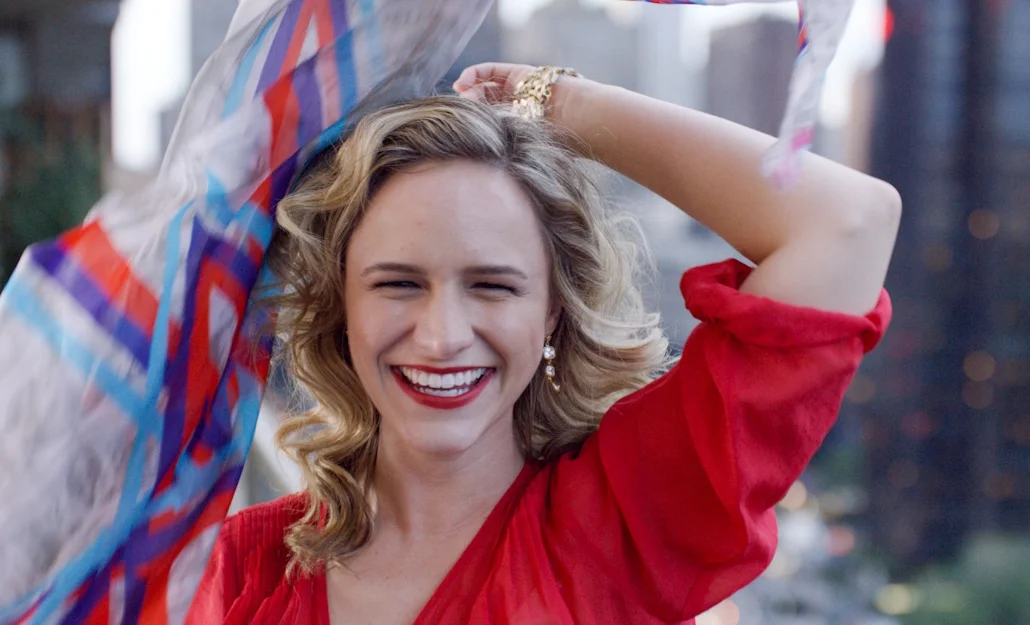 A woman with blonde curly hair wearing a red dress is smiling and holding colorful shopping bags, standing outdoors.