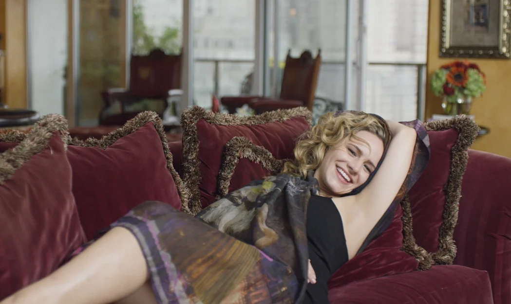 A woman with blonde curly hair lying on a red velvet sofa, smiling and stretching with one arm behind her head, in a cozy room with large windows, chairs, and a vase of flowers in the background.