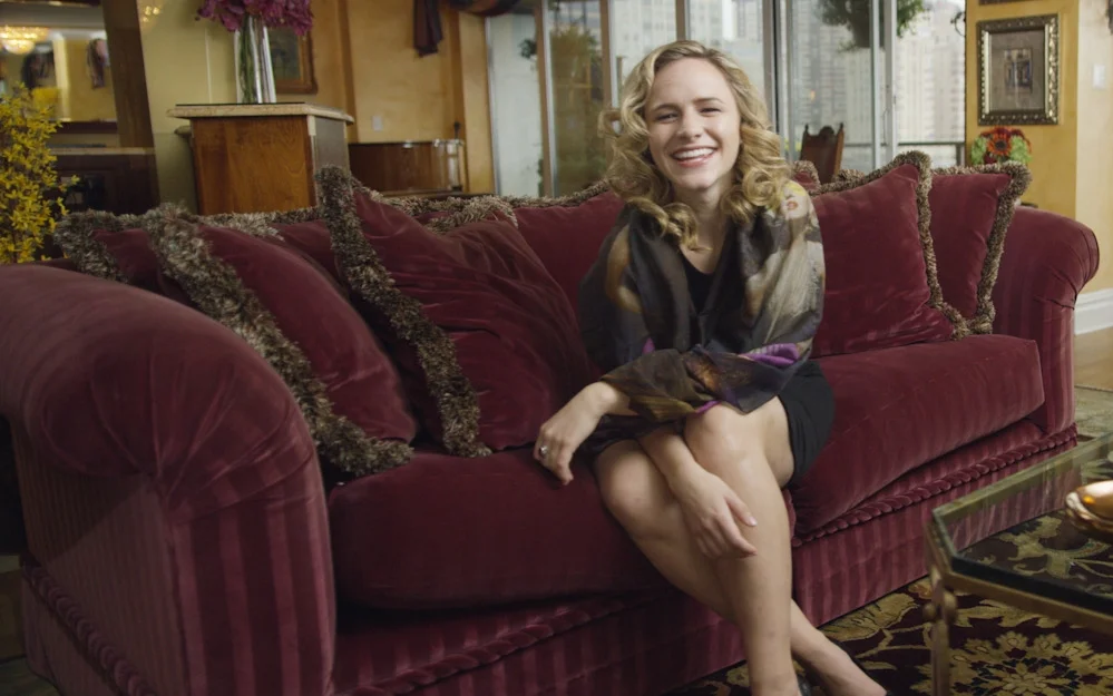 A woman with curly blonde hair smiling while sitting on a red velvet sofa in a warmly decorated living room.