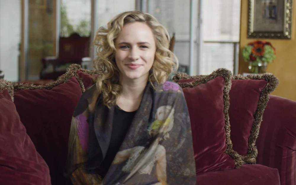 A woman with blonde curly hair sitting on a red couch with a patterned shawl, in a warmly decorated room with flowers and artwork.