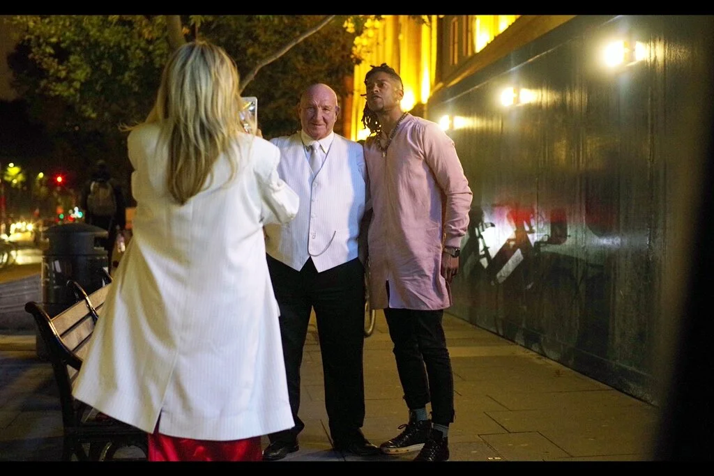 “Darling, take a photo of me standing next to this young rapper.” ”Actually, I’m a practising endocrinologist.” ”Maybe, but when I update my Facebook profile picture you won’t be” The man in the white vest is in this movie - he’s Dave Courtney.