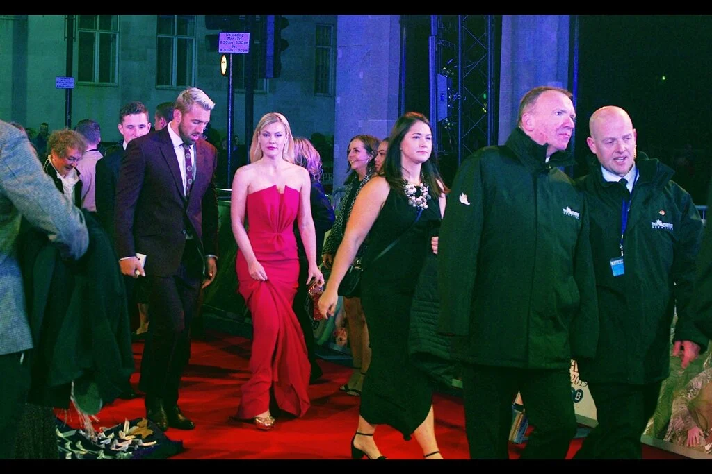 Camilla Kerslake seems ambivalent about the difficulty of correcting the white balance in this photo when everything is/was mostly red, including the ACTUAL red carpet and her dress. Security on the right appear to be concerned about something, but sadly I’m certain it’s not about the difficulty of establishing white balance in this environment.