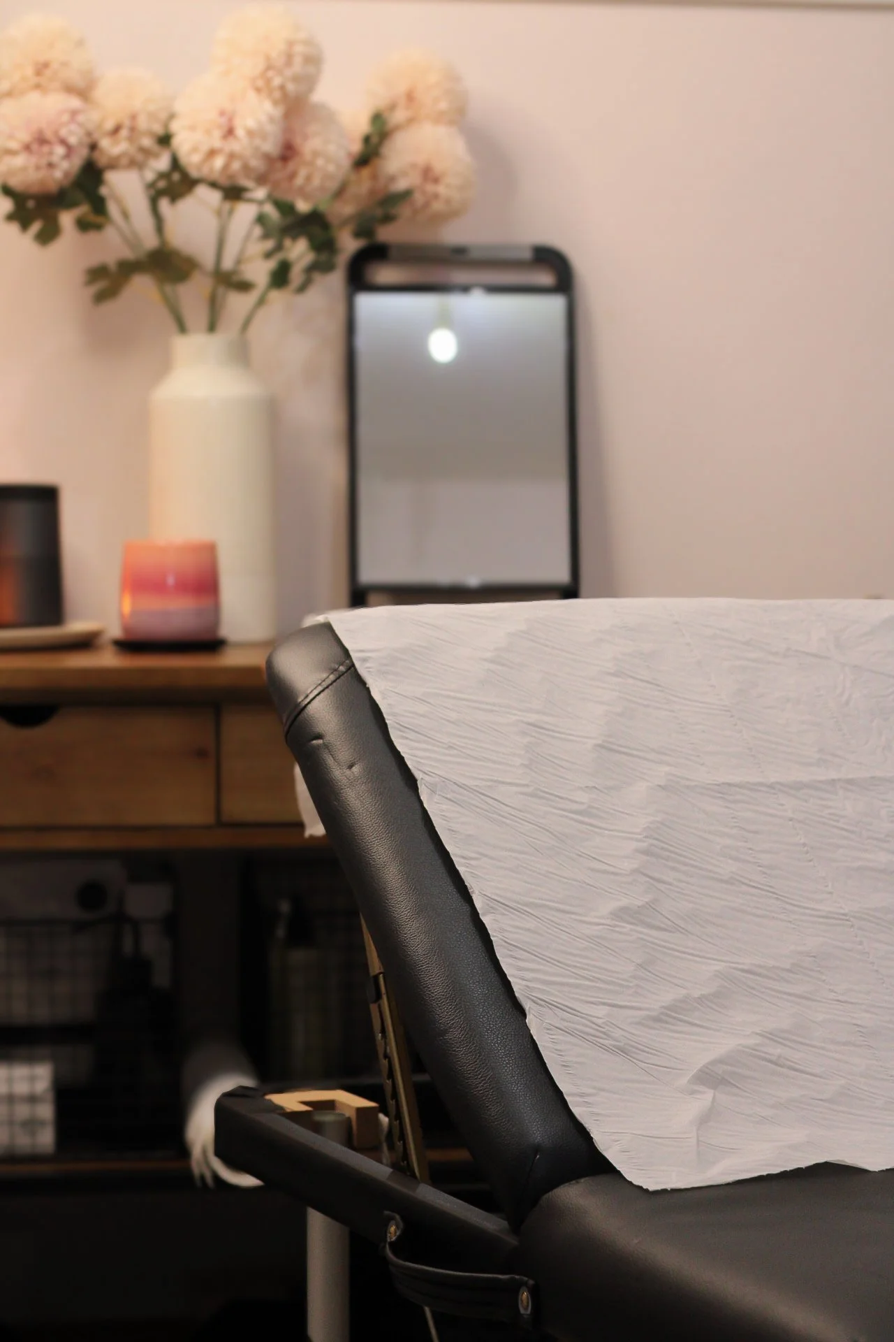 Treatment bed with paper sheet, wooden table with flower vase, candle, mirror, and storage baskets in the background.