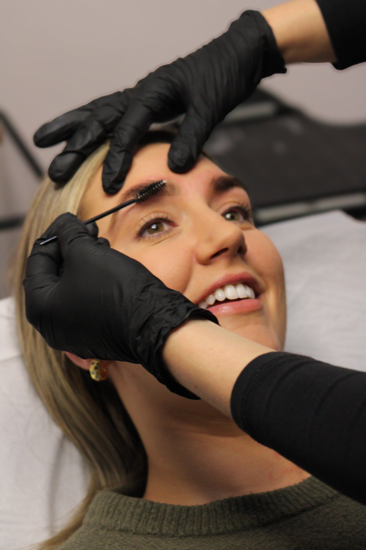 A woman getting her eyebrows styled by a professional wearing black gloves, who is applying a cosmetic product with a wand to her eyebrows.