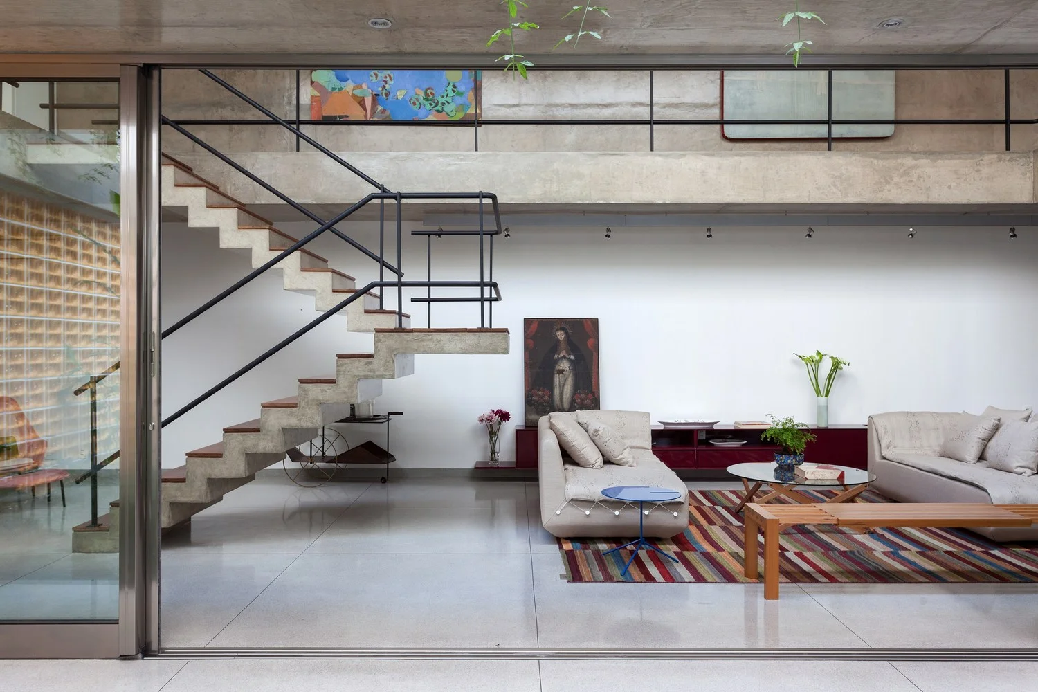 Pé-direito duplo da sala de estar com piso e escada em concreto aparente e piso de granilite. Ao fundo, trilho de iluminação. À esquerda, cobogó amarelo.