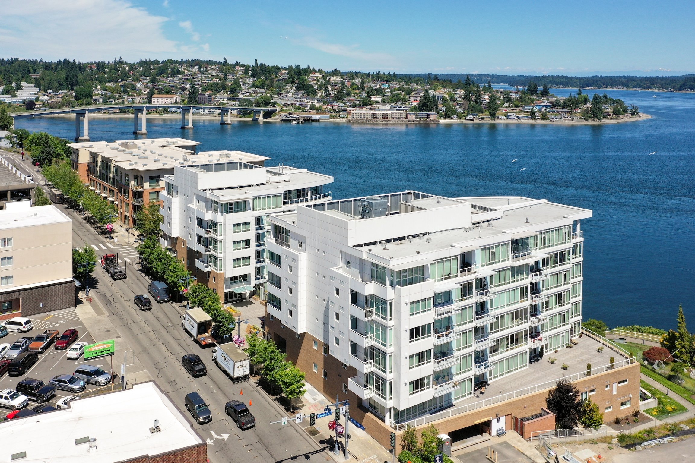 Aerial view of modern apartment buildings along a waterfront, with a bridge and lush greenery in the background under a clear blue sky.