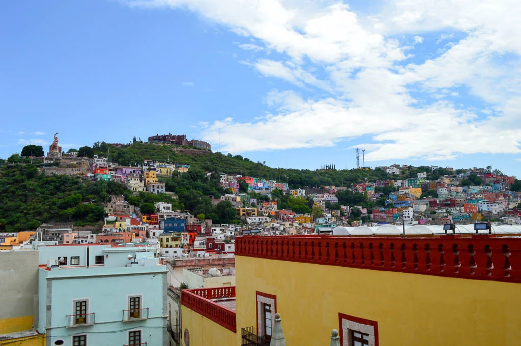 View across the colorful houses of Guanajuato, Mexico, with the El Pípila monument on the hillside above the city and a yellow and red colonial building in the foreground.