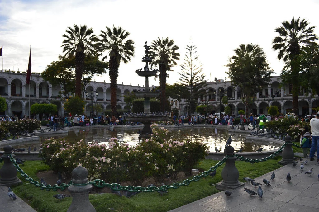 A view of the Plaza de Armas of the historic district of Arequipa Peru at sunset. It is a view of the fountain at sunset with families and pigeons sitting around the fountain.
