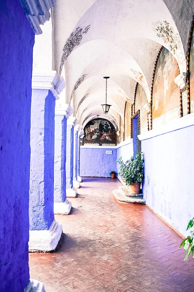 A view of a passageway in Monasterio de Santa Catalina in Arequipa Peru. The ceilings are arched in white sillar and the walls and columns are blue.