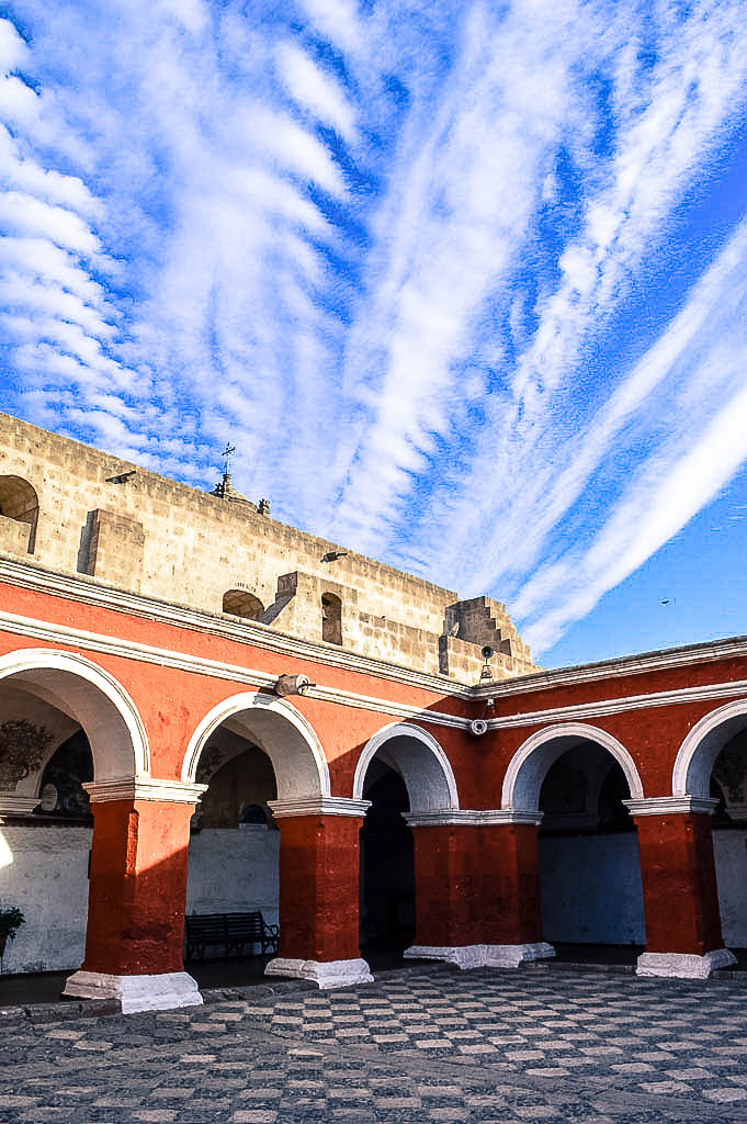 A view of a courtyard in the Monasterio de Santa Catalina in Arequipa Peru. The sky is visible. The walls are arched and red and the floors are black and white cobbles.