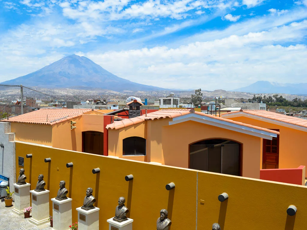 View of the volcano at the Mirador de Yanahuarra viewpoint. There are brightly colored orange houses, with a yellow wall, with busts of important figures in Peru.