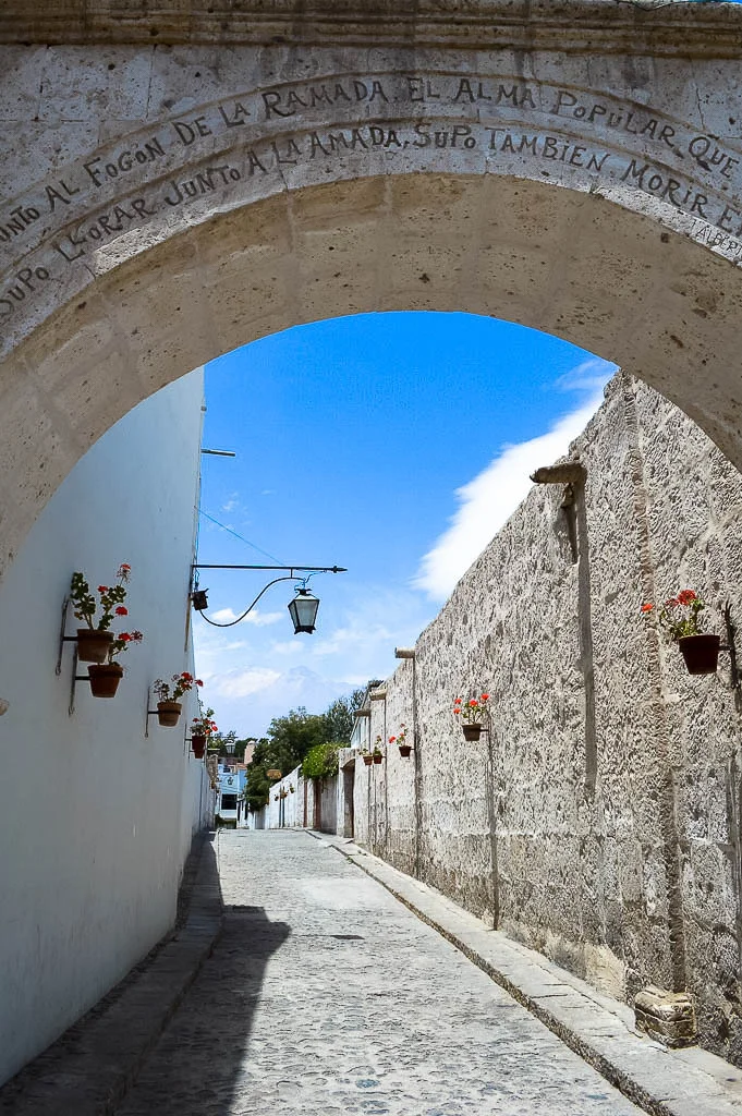 A alleyway made of white sillar wall with pot plants hanging from it. The volcano is visible in the background in Arequipa Peru.