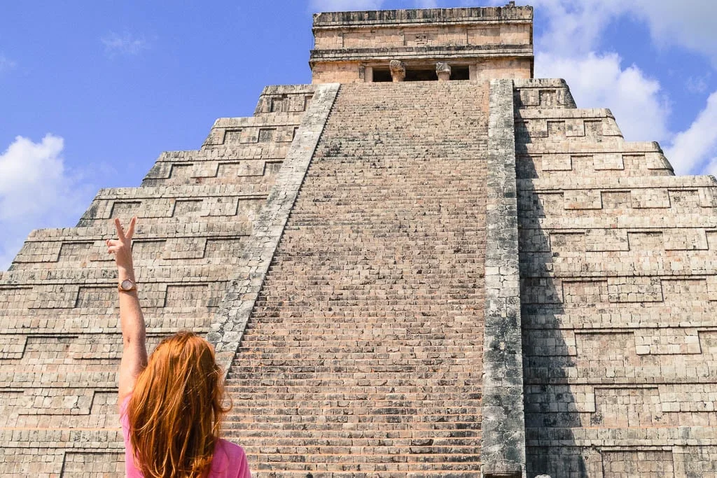 A red headed woman in a pink top holding up a peace sign in front of the El Castillo pyramid at Chichén Itzá, the Mayan ruins in Mexico's Yucatán peninsula.