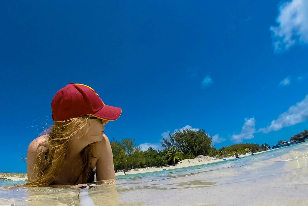A red headed woman in a red baseball cap lying in the shallow clear water at the edge of a white sand beach in The Bahamas.