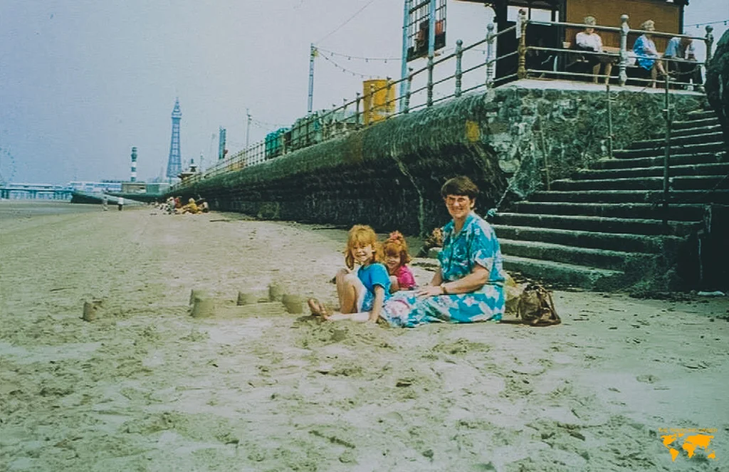 Two red headed children and a grandmother sitting on the beach building sandcastles in Blackpool with the Blackpool tower in the distance.
