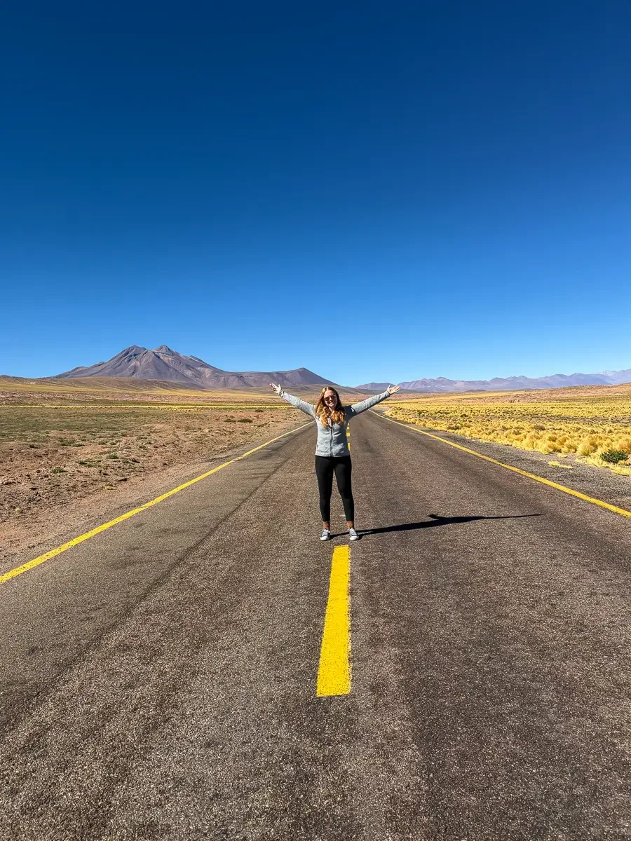 Woman standing with arms outstretched in the middle of an empty desert highway with volcanoes ahead, on the self-drive route to Lagunas Miscanti, Chile