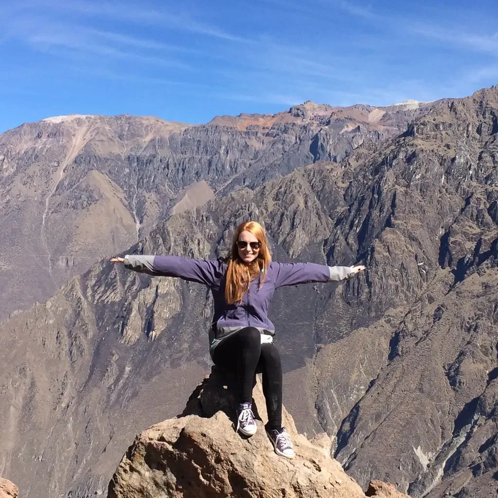 Red head woman sitting on a rock overlooking Colca Canyon with arms spread wide like a condor, dressed in purple.