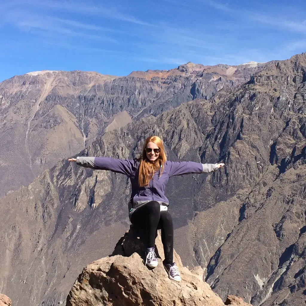 Visiting Colca Canyo. Red head woman sitting on a rock overlooking Colca Canyon with arms spread wide like a condor, dressed in purple.