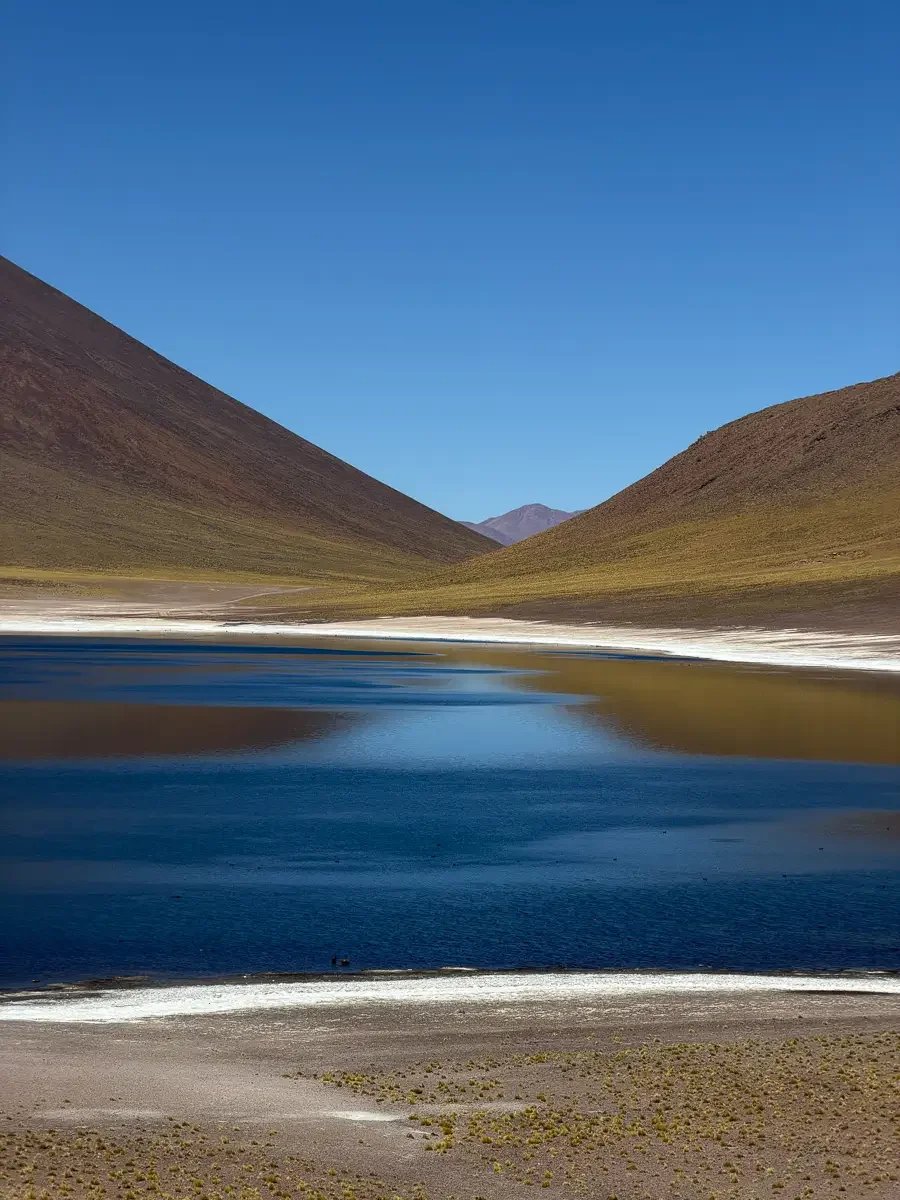 Deep blue water of Laguna Miscanti edged with white salt crust, framed by two volcanic hillsides with a distant peak visible between them, Atacama Desert, Chile