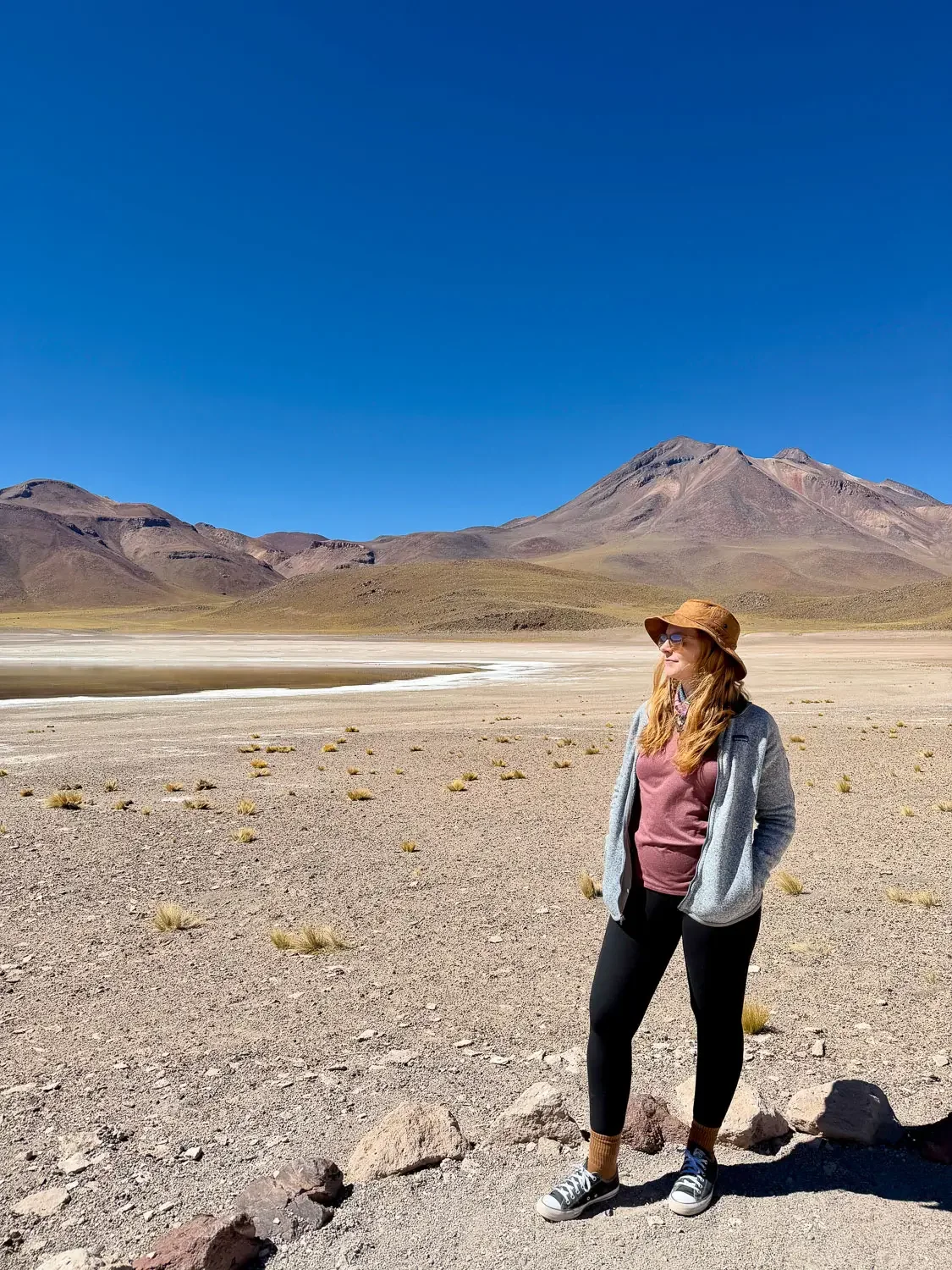 Woman in a fleece jacket, leggings, sneakers and sun hat standing on the gravel shore of an altiplanic lagoon with volcanoes behind, Atacama Desert, Chile