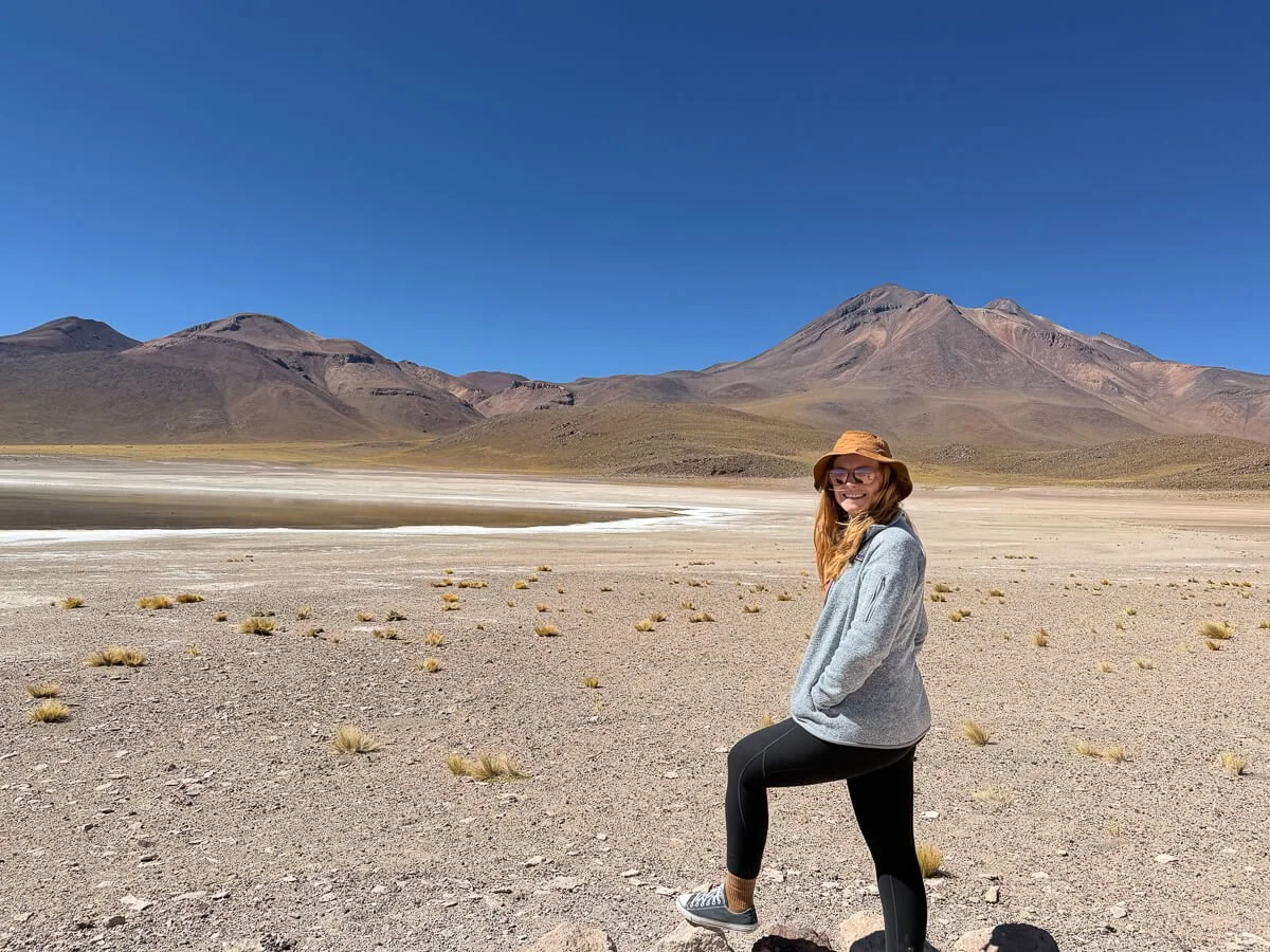 Woman in a fleece and sun hat on the gravel shore of an altiplanic lagoon with volcanic peaks behind, Los Flamencos National Reserve, Chile