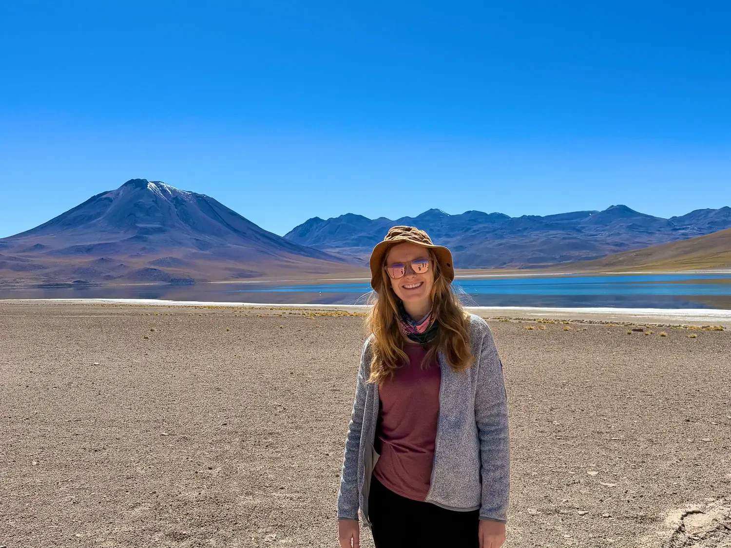 Laguna Miscanti in Chile's Atacama Desert, with a snow-dusted volcano behind the bright blue water and a woman in a fleece and sun hat standing on the gravel shore