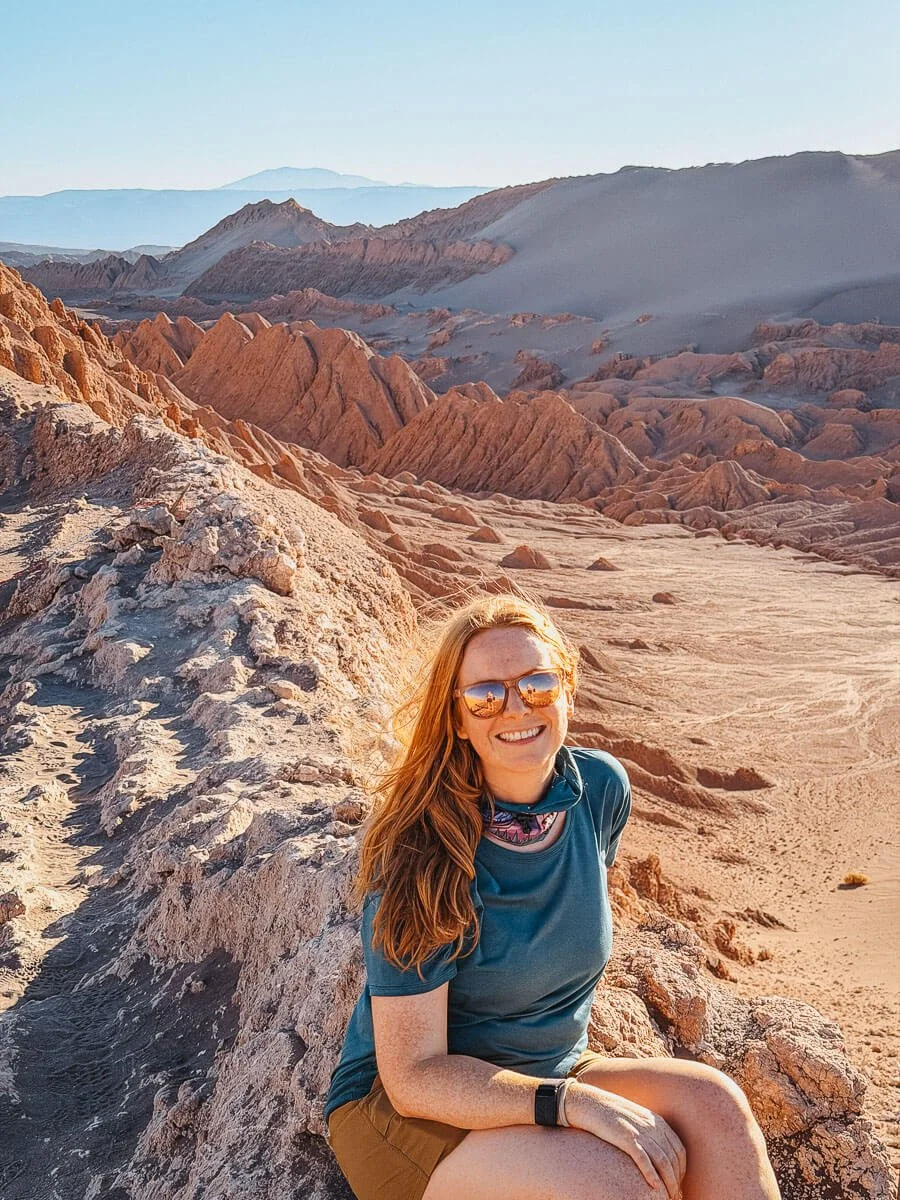 Ginger girl in hiking gear sitting on rocks infront of rock formations next to the duna mayor in valle de la luna in the atacama desert