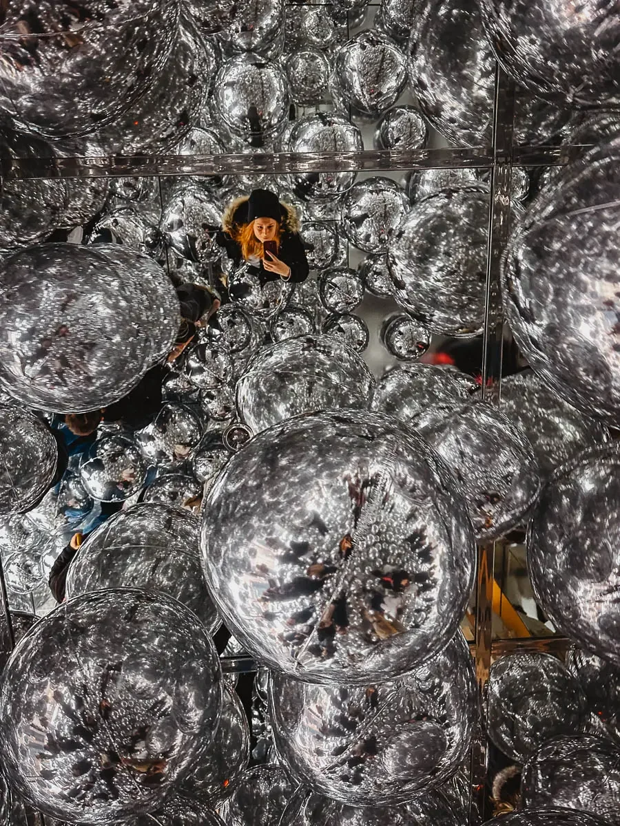 art exhibition of bright silver round balls floating around the room with a red headed woman taking a selfie amongst them at the summit one vanderbilt