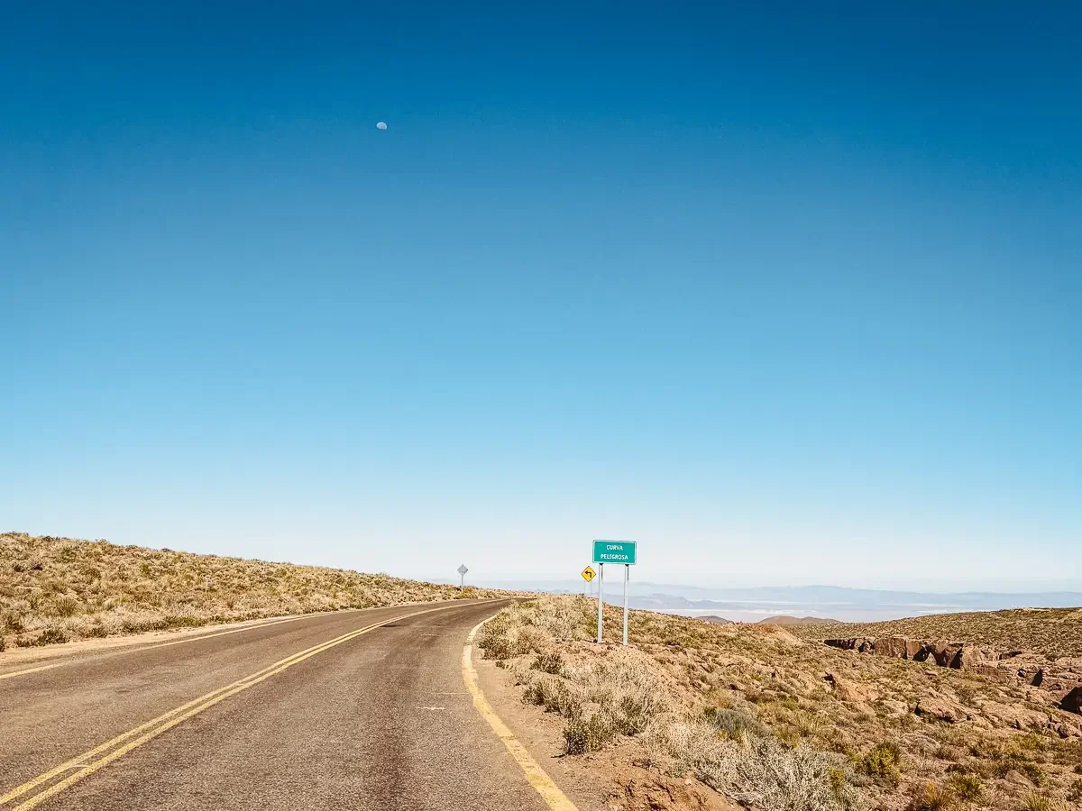 Empty paved highway curving through the high desert on the drive to Lagunas Miscanti, with a dangerous curve sign and the moon in a clear blue sky, Atacama Desert, Chile