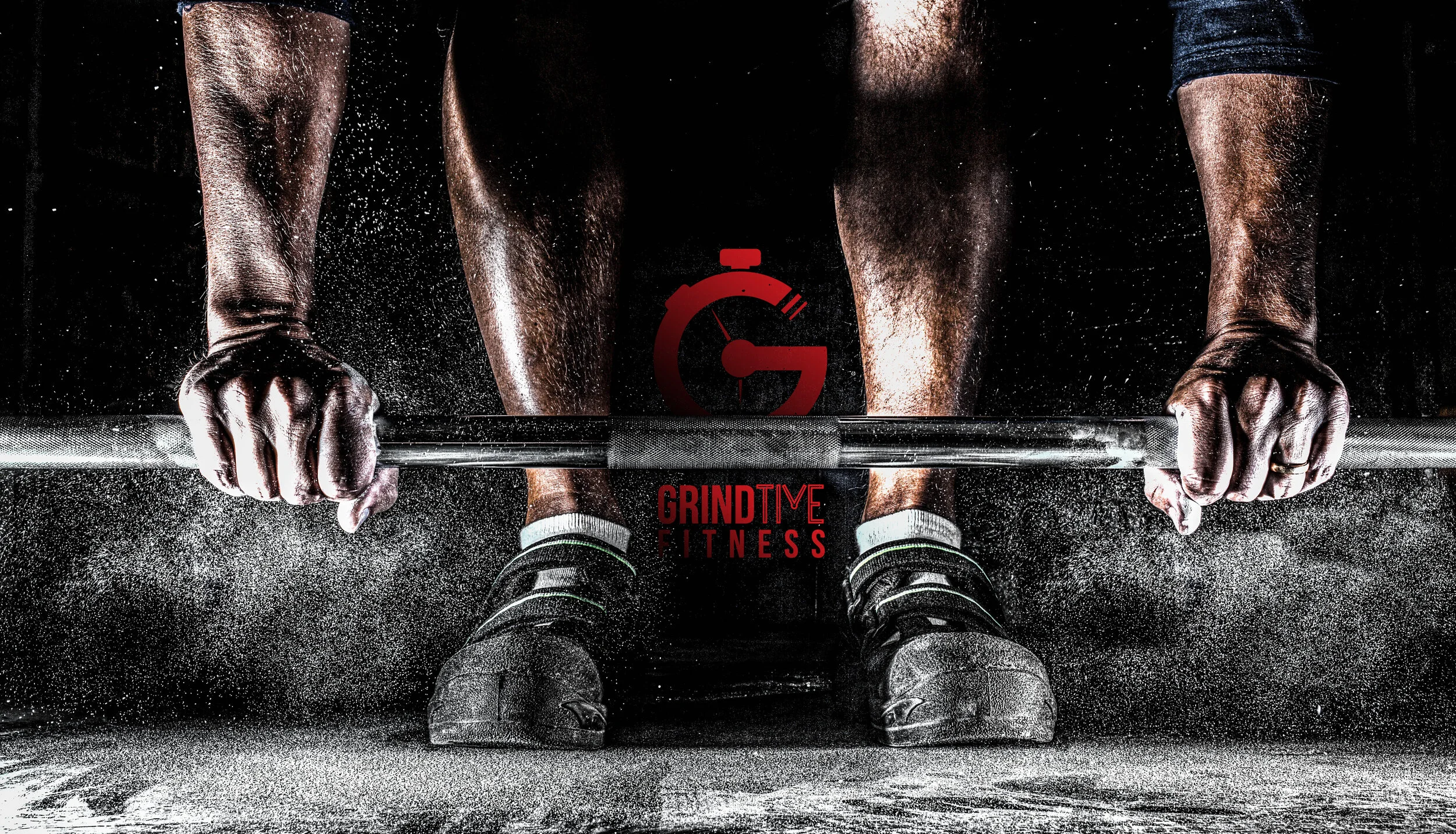 Close-up of a person performing a deadlift exercise, with hands gripping a barbell, covered in chalk dust, in a dimly lit gym with fitness branding in the background.