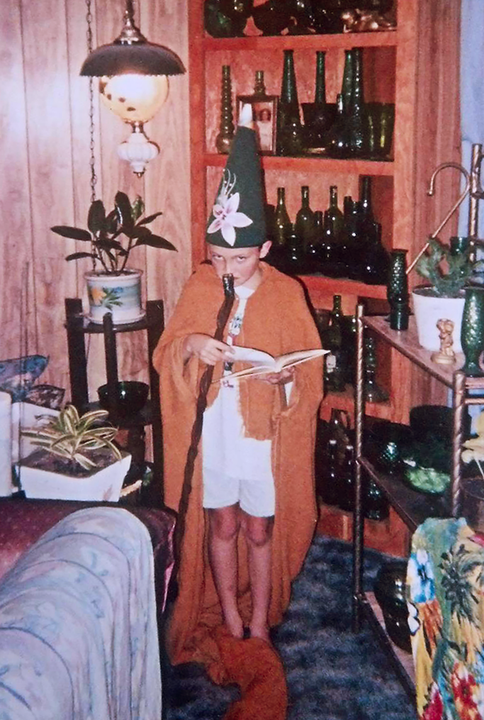 Child wearing a magician's hat with a flower, a long cape, and holding a wand, standing in a cozy wood-paneled room with plants, shelves, and furniture.
