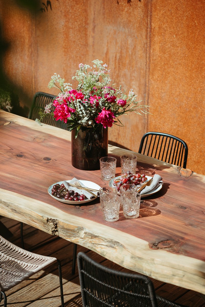 Ein rustikaler Holztisch mit einer großen Vase voller pinkfarbener und weißer Blumen, umgeben von Gläsern, Geschirr und Weintrauben vor einer orangefarbenen Wand.