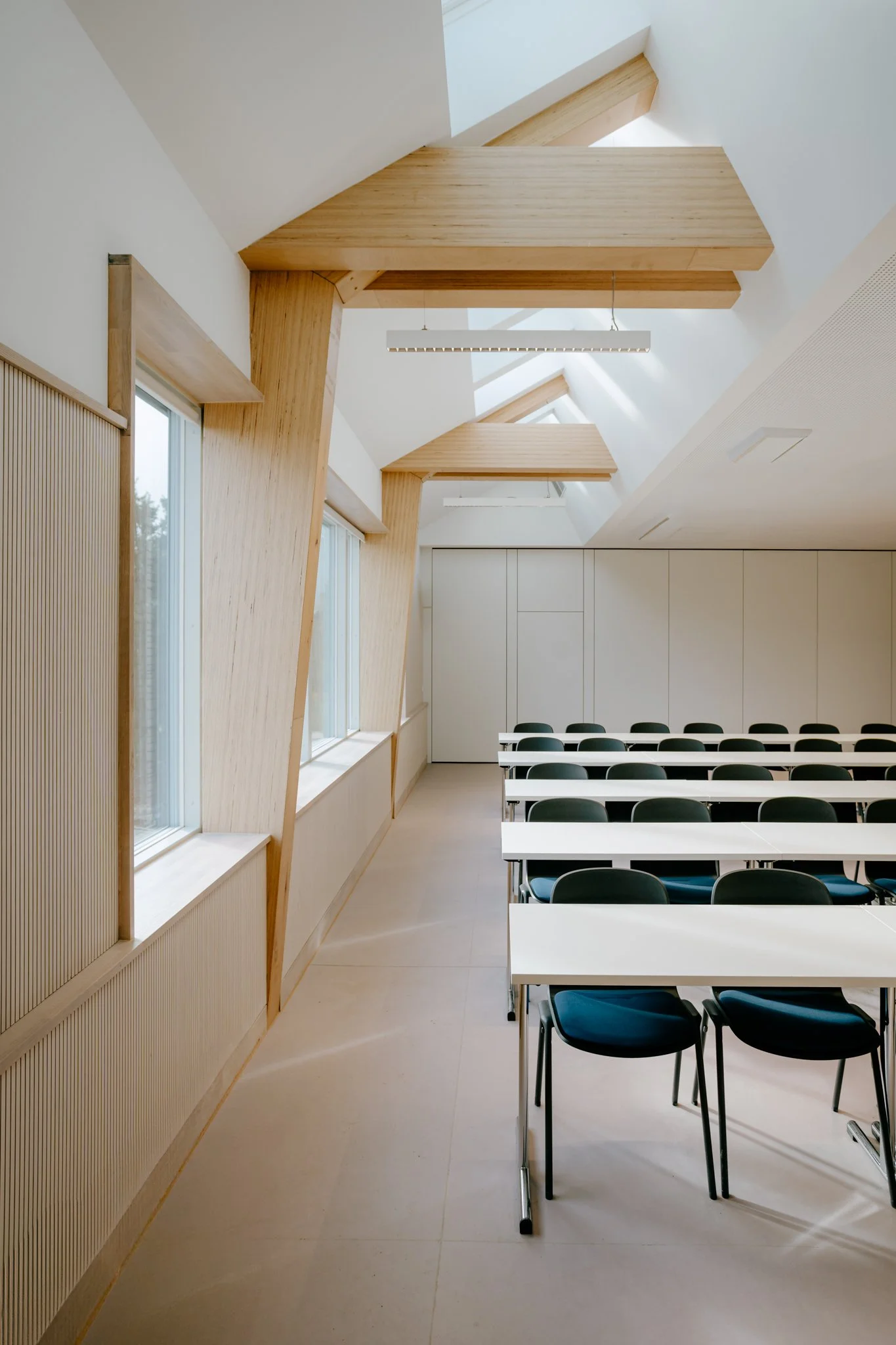 Innenraum eines modernen, hellen Konferenzraums mit weißen Tischen, schwarzen Stühlen und großen Fenstern, Decke mit Holzpaneelen und Dachfenstern.