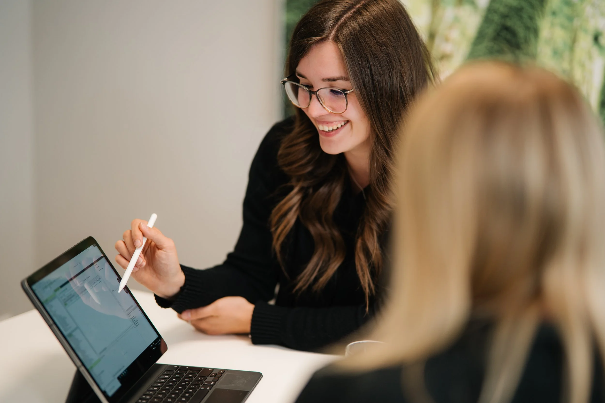 Eine junge Frau mit Brille und braunen, gewellten Haaren, die auf einem Laptop arbeitet und mit einer anderen Person spricht, die nur teilweise im Bild sichtbar ist.