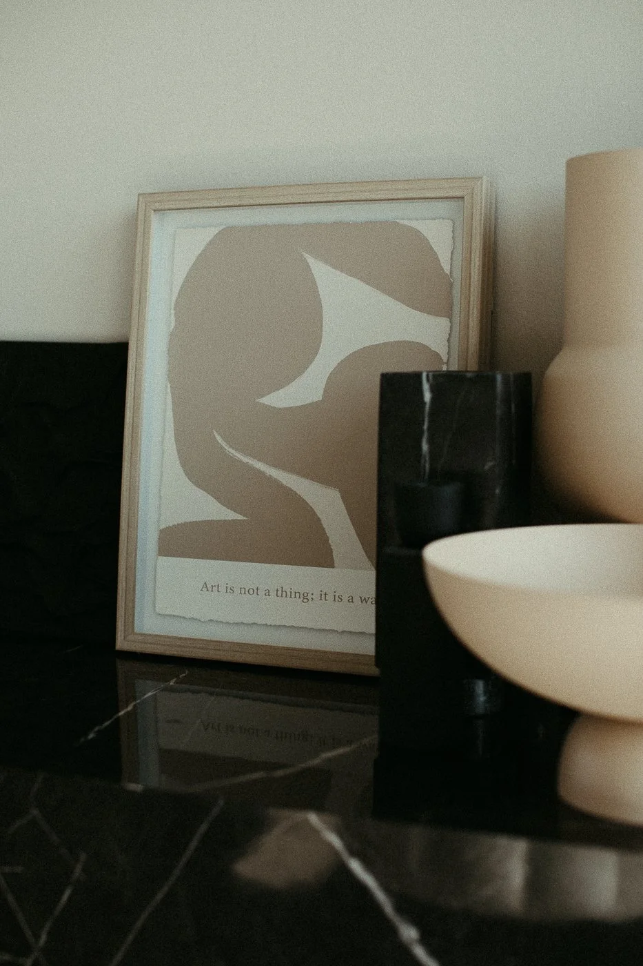 Ein eingerahmtes Kunstwerk mit abstrakten Formen und einem Zitat, das auf einem schwarzen Marmortisch steht, neben einer schwarzen Vase und einer beige Vase.