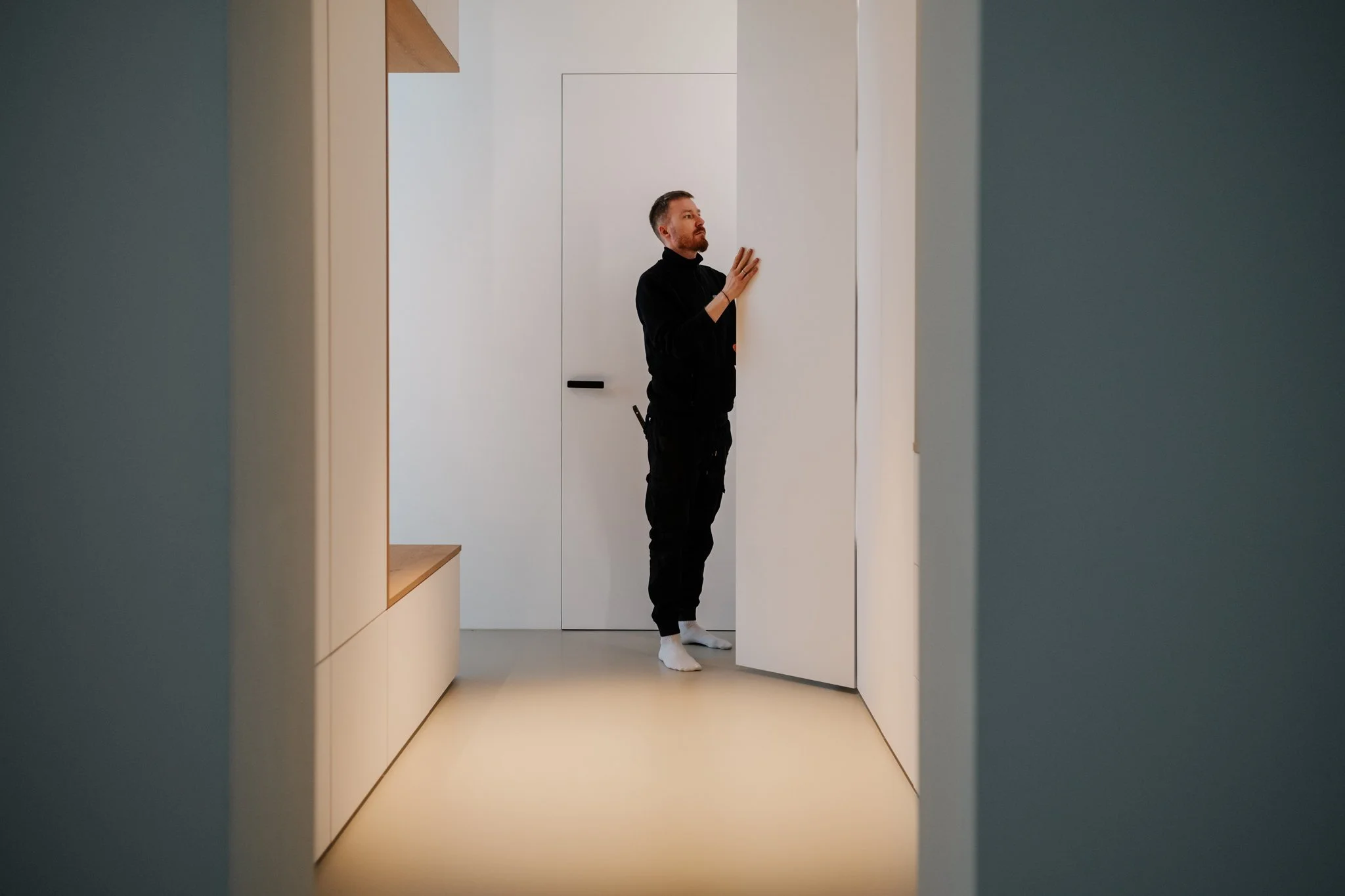 Ein Mann steht in einem minimalistischen, weißen Raum mit weißen Wänden und Türen, während er eine Tür schließt oder öffnet.