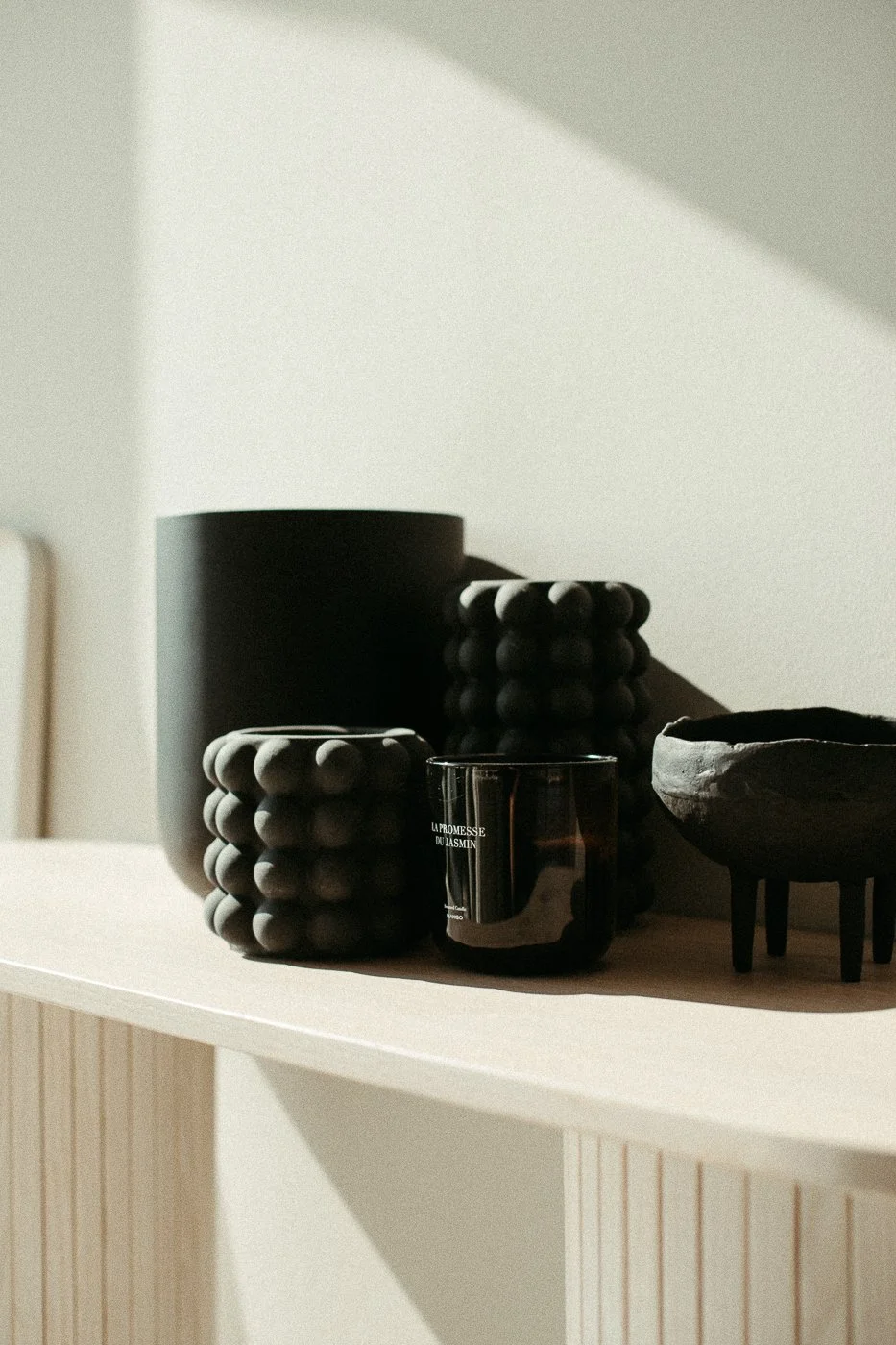 Schwarz-glänzende Kerze mit weißen Buchstaben, schwarze dekorative Gegenstände mit Ballformen, eine schwarze Schale auf einem hellen Regal, Schatten an der Wand.