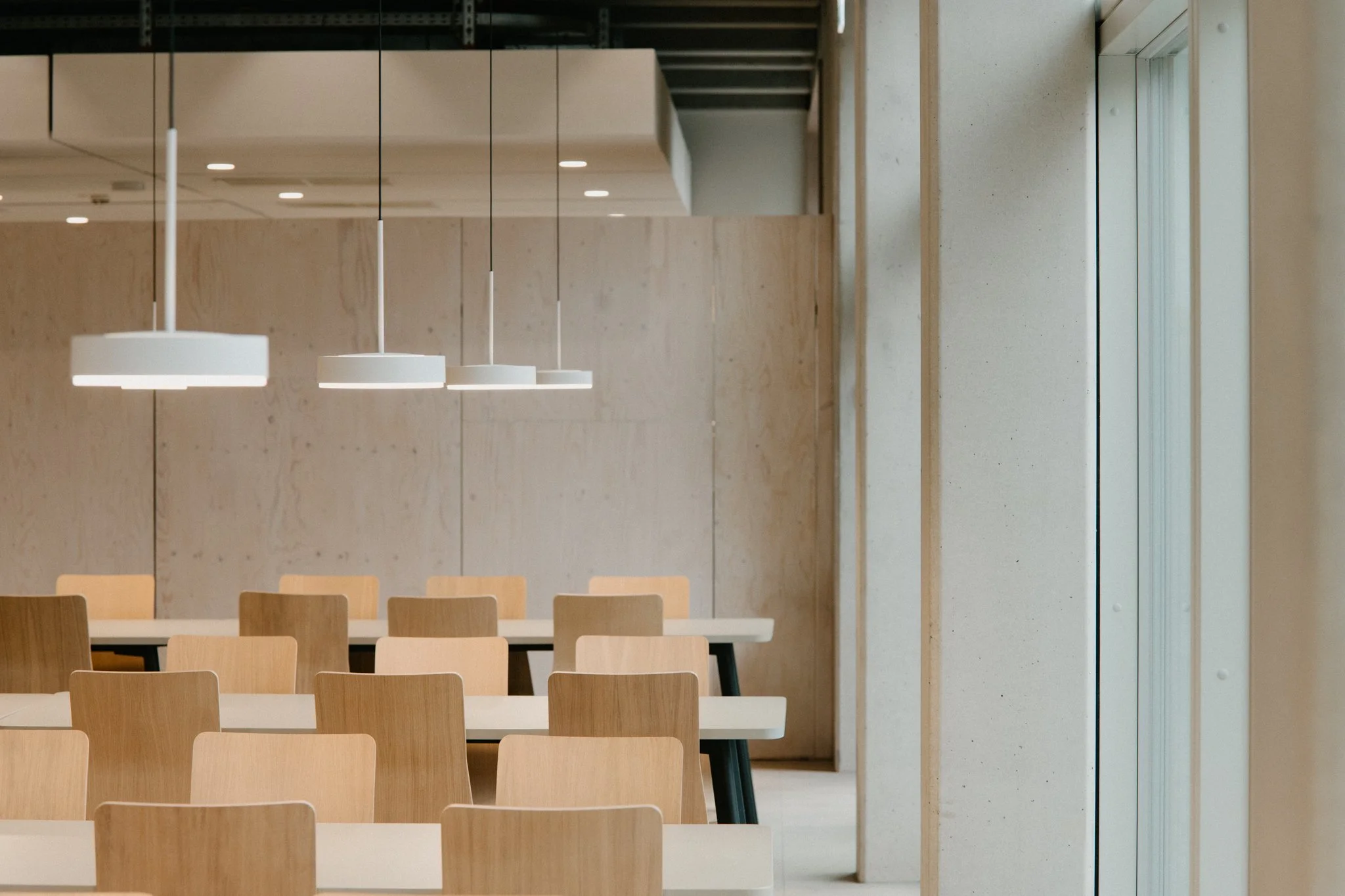 Innenraum eines modernen, minimalistischen Raumes mit Holzmöbeln, weißen Tischen und weißen Hängelampen, natürliche Lichtquelle durch die Fenster.