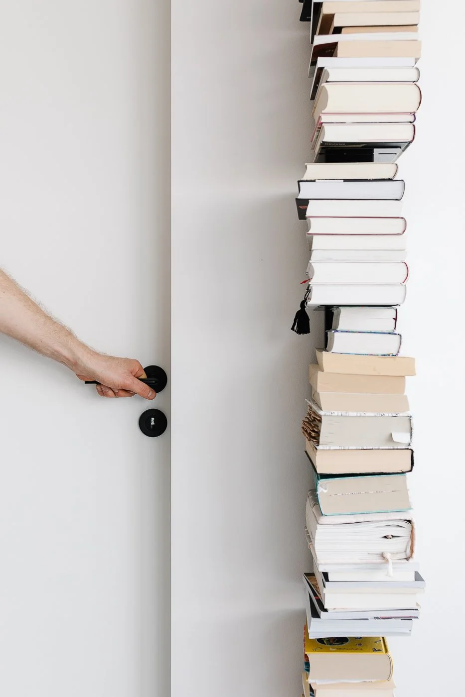 Ein weißer Raum mit einer Hand, die eine graue Tür mit einem schwarzen Knauf öffnet, neben einer hohen Turm aus Büchern.
