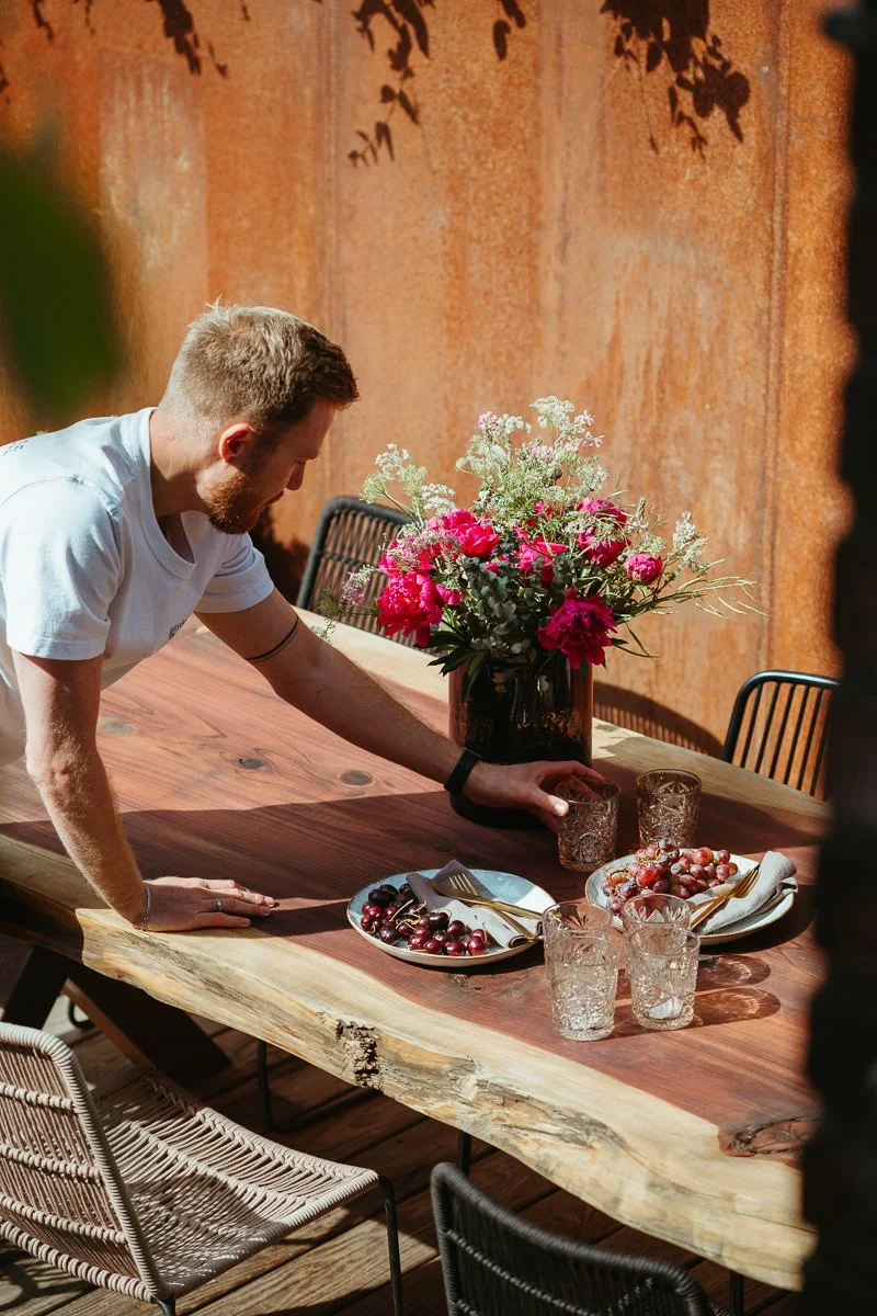 Ein Mann bereitet einen Tisch mit frischen Kirschen, Gläsern und Besteck für eine Mahlzeit vor, auf einem Tisch mit Blumen im Hintergrund.
